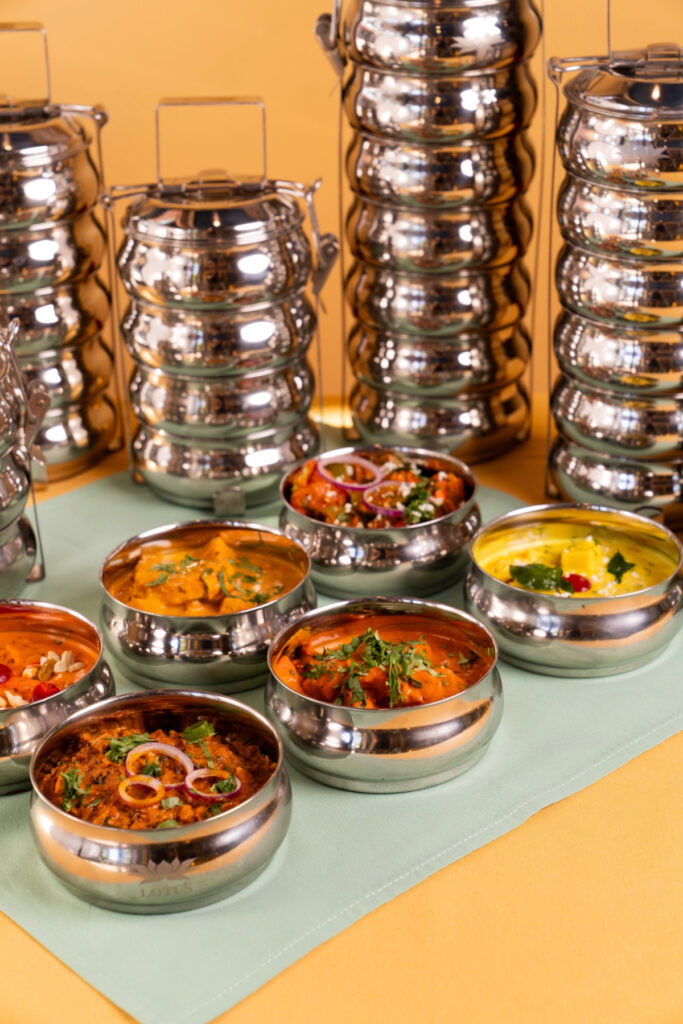 A colorful spread of Indian dishes served in shiny stainless steel bowls with stacked tiffin containers in the background, showcasing Lotus Cuisine of India’s reusable takeout system.