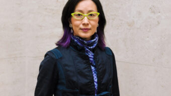 Dr. Soyoung Lee, director of the Asian Art Museum, wearing lime green glasses and a dark jacket with purple accents, standing against a textured wall.