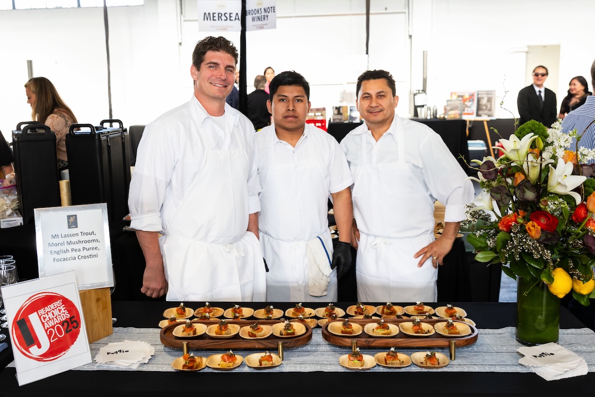 Three chefs in clean white uniforms stand behind a table with plates of food.