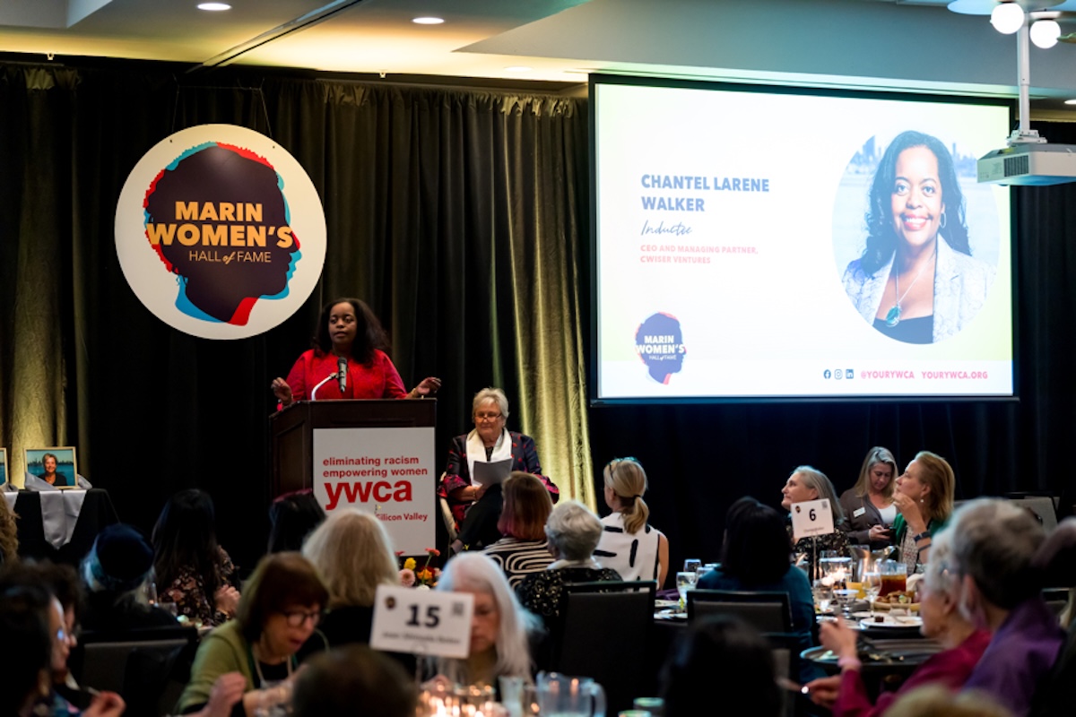 A woman stands behind a podium making an acceptance speech, with a big circular white poster with a graphic of a silhouette head and the words "Marin Women's Hall of Fame" and a screen with a slider and a photo of the woman.