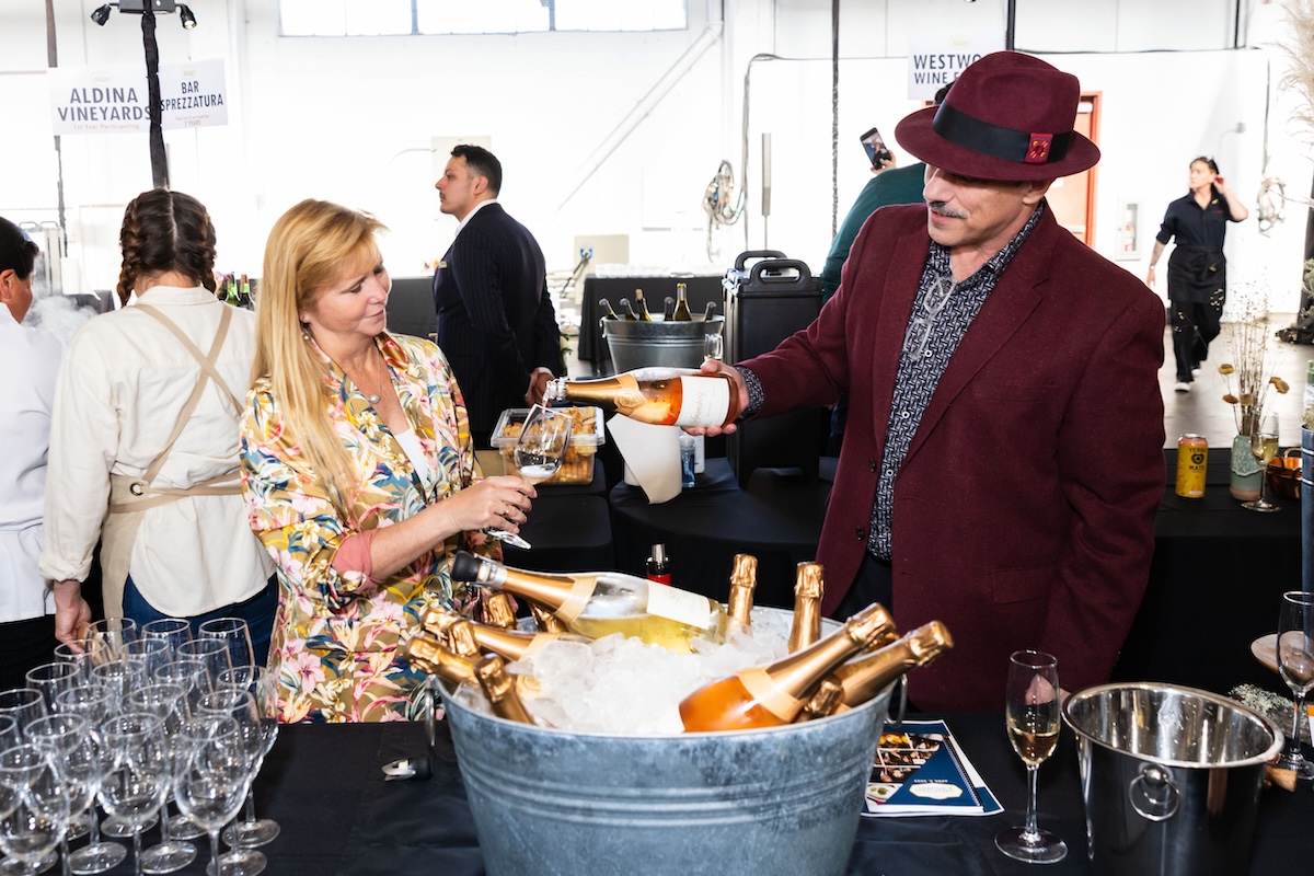A man in a burgundy suit and fedora pours wine from a bottle into a woman's glass.