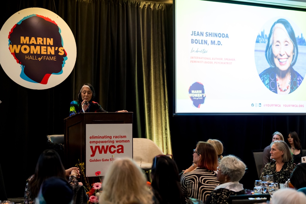 A woman stands behind a podium making an acceptance speech, with a big circular white poster with a graphic of a silhouette head and the words "Marin Women's Hall of Fame" and a screen with a slider and a photo of the woman.