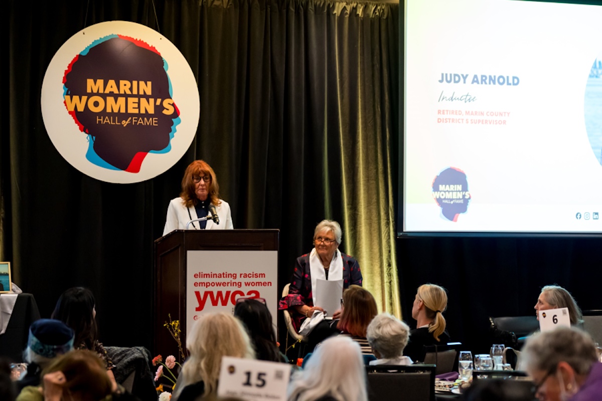 A woman stands behind a podium making an acceptance speech, with a big circular white poster with a graphic of a silhouette head and the words "Marin Women's Hall of Fame" and a screen with a slider and a photo of the woman.