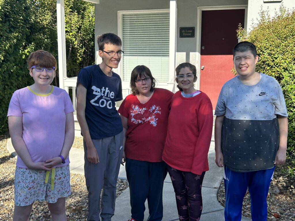 A group of five young adults stands in front of a house with a red front door.