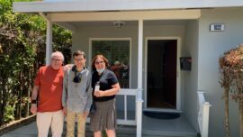 Two men and a woman stand in front of a house with a front porch.