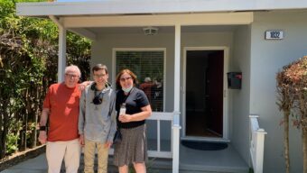 Two men and a woman stand in front of a house with a front porch.