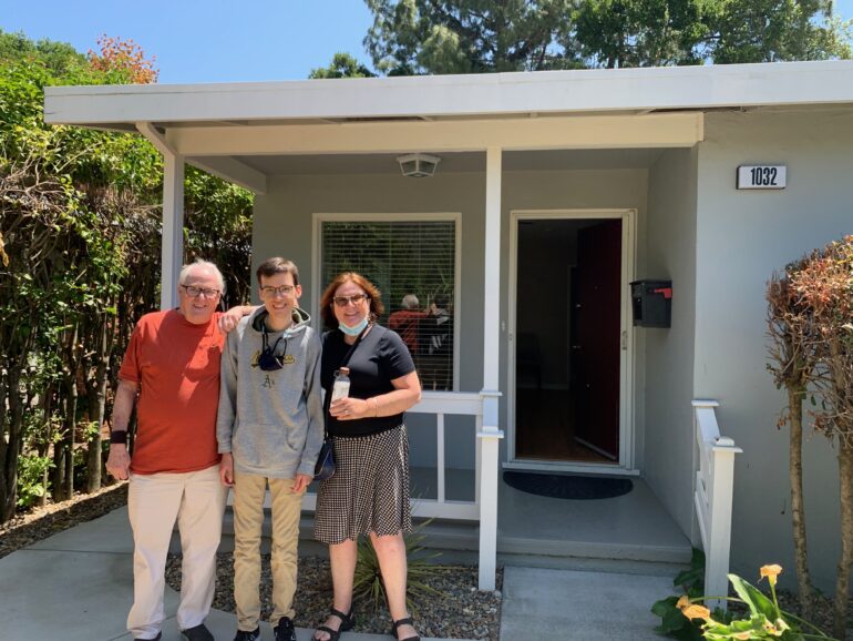 Two men and a woman stand in front of a house with a front porch.