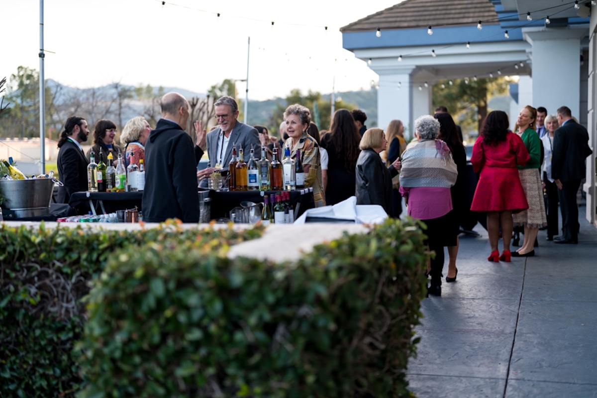 A hedge with a view of a party as people walk around and visit an outdoor bar.