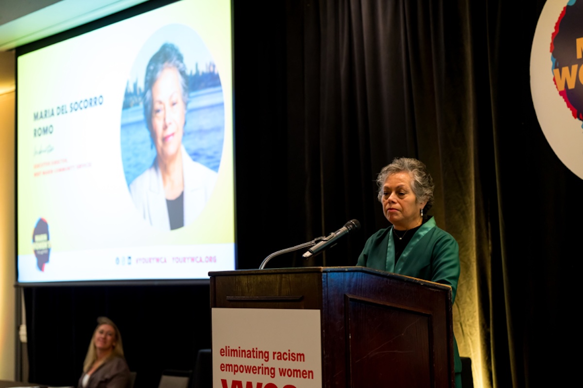 A woman wearing dark greeen stands behind a podium making an acceptance speech, with a big screen showing a slide with her photo and name.