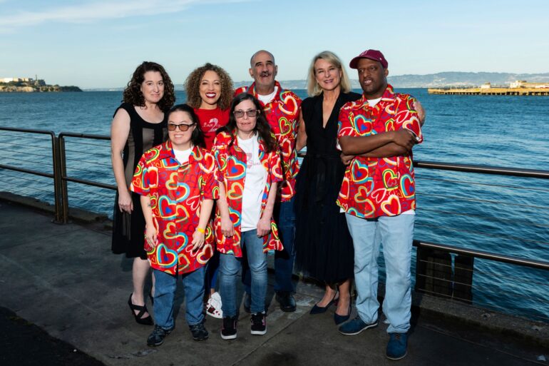A group of seven people, including four wearing matching red shirts with rainbow hearts, stand on a deck right next to the bay.