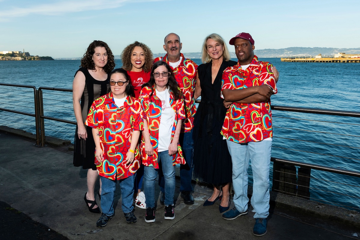 A group of seven people, including four wearing matching red shirts with rainbow hearts, stand on a deck right next to the bay.