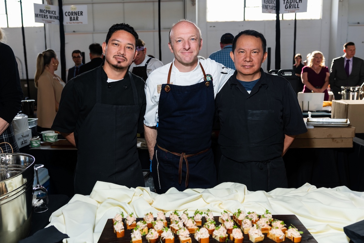 Three chefs in black chef attire stand behind a table with bite-sized food on a tray.