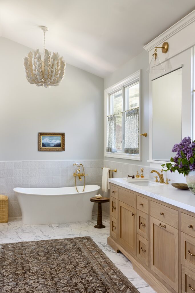 Serene bathroom with a freestanding soaking tub, gold fixtures, a natural wood vanity, marble flooring, and a dramatic white petal chandelier under vaulted ceilings.