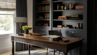 Home office with a rustic wooden desk, cane-back chair, dark built-in bookshelves, and a large window dressed with textured Roman shades, creating a cozy and functional workspace.