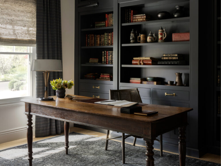 Home office with a rustic wooden desk, cane-back chair, dark built-in bookshelves, and a large window dressed with textured Roman shades, creating a cozy and functional workspace.