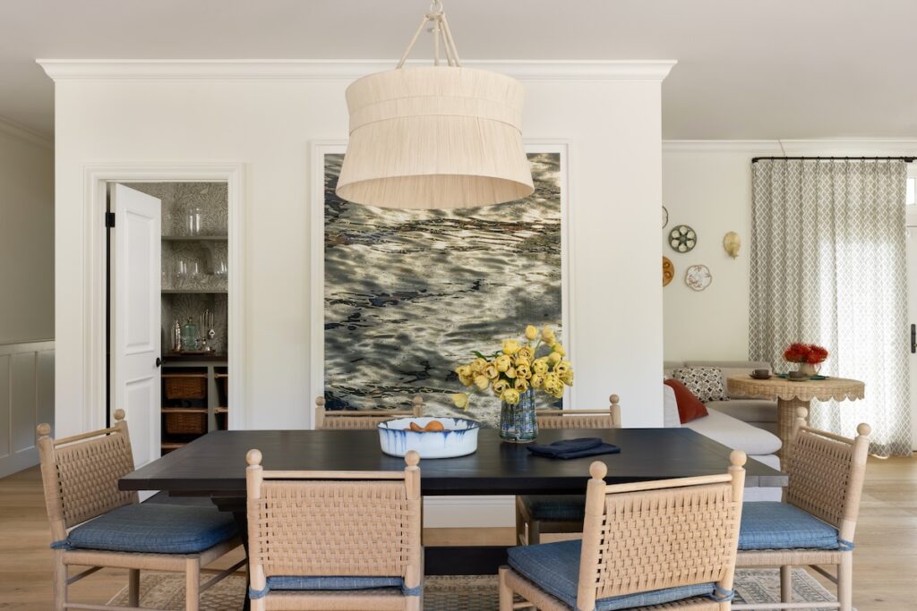 Open-concept dining area with a dark-stained Elsie Green dining table, woven oak Le Maé chairs, oversized Palecek pendant, and a large water-themed photograph by Barbara Vaughn, adjacent to a cozy breakfast nook.