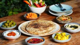 Centered on the table is a plate with pita bread, and there are bowls of beet dip, hummus, fruit, olives, carrots and more around it.