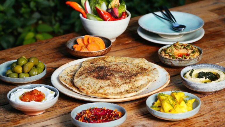 Centered on the table is a plate with pita bread, and there are bowls of beet dip, hummus, fruit, olives, carrots and more around it.