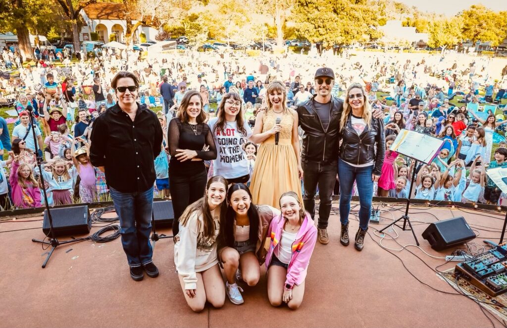A group of nine people pose for an image on a stage with a large crowd behind them.