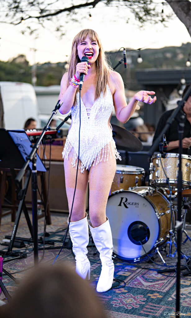 A young woman with blonde hair and bangs and wearing a white sparkly bodysuit and white knee-high boots sings on stage.