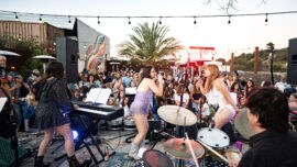 Two women sing on stage wearing sparkly bodysuits while a band plays behind them and a large crowd watches.