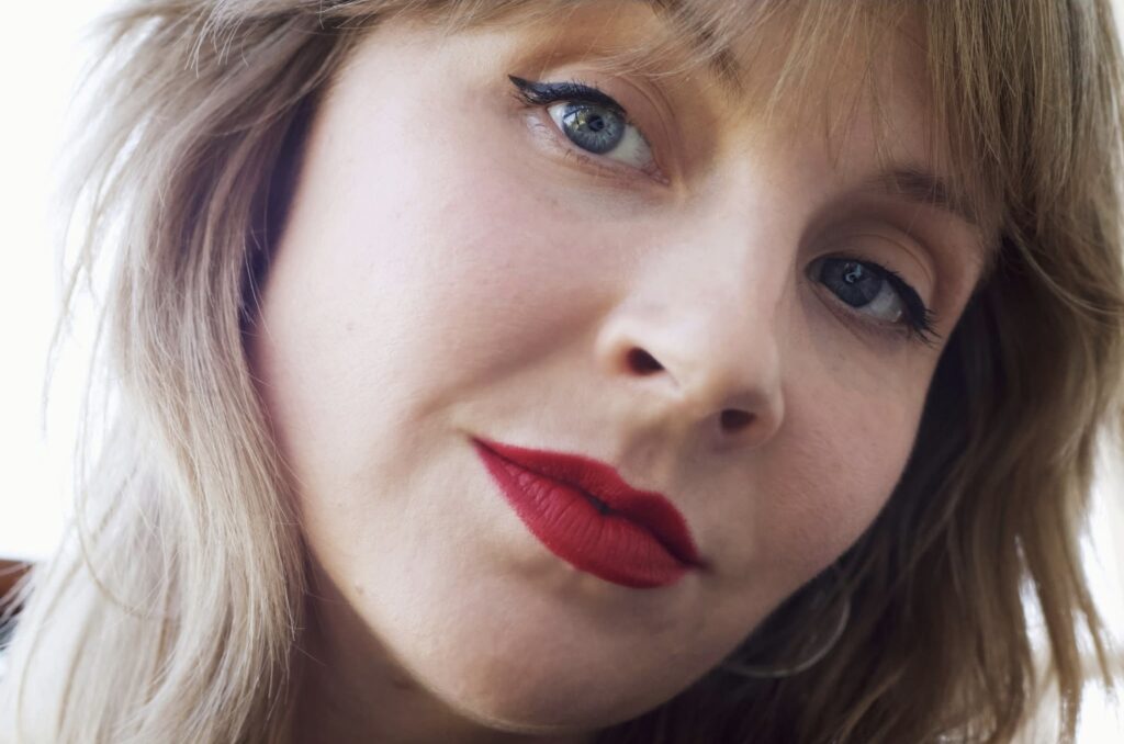 A young woman with blonde hair, bangs, red lipstick and winged eyeliner.