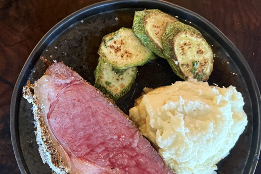 A plate with a slice of rare steak and some sliced veggies and mashed potatoes.