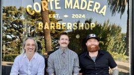 Three men sit outside a window with the words "Corte Madera Barbershop."