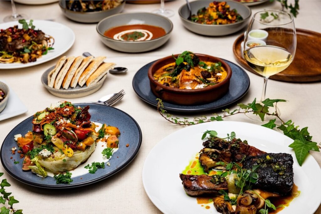 A table set with many plates of intricately plated meals and some glasses of white wine.