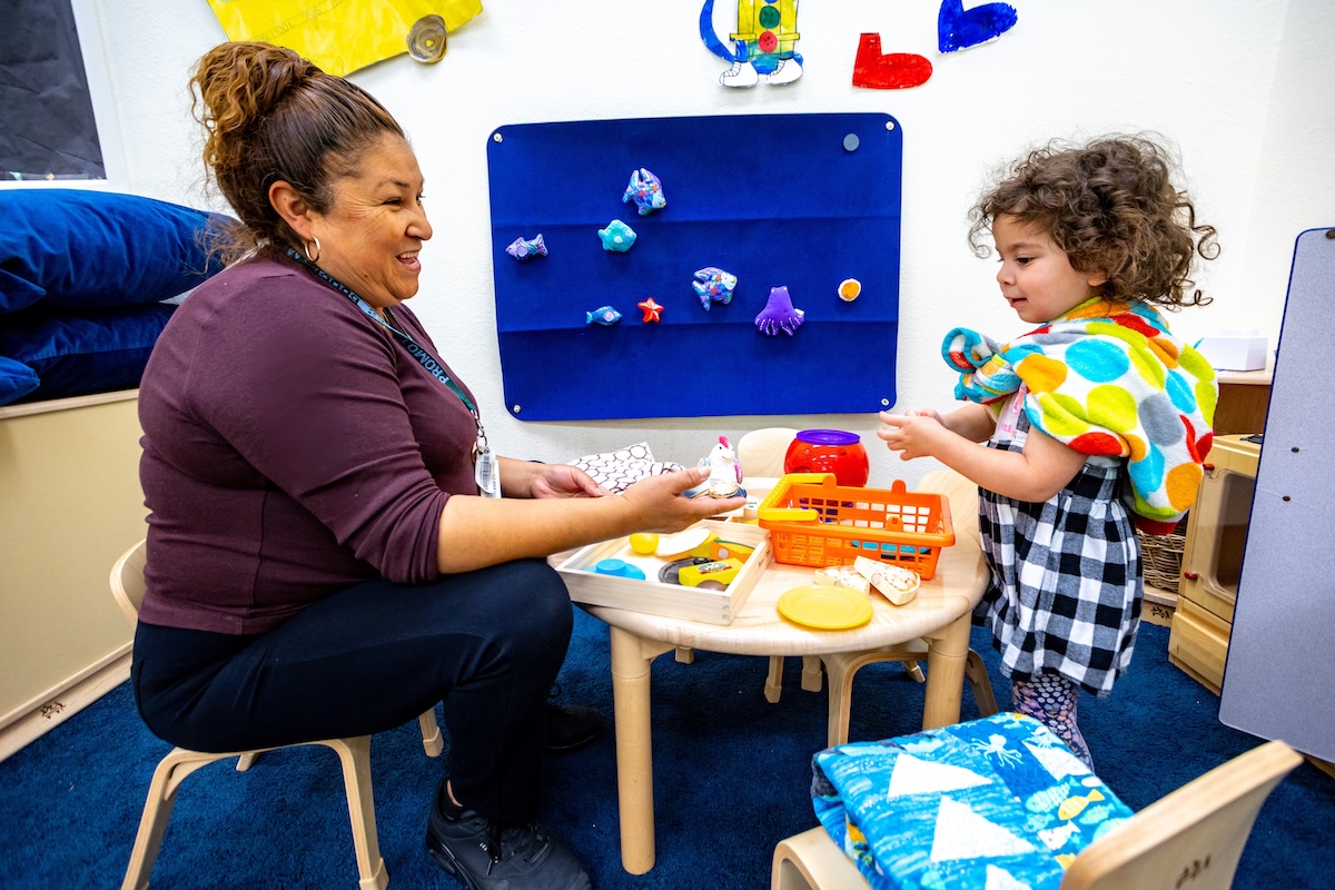 A woman plays with a kid at a small table with lots of toys.