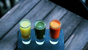 Three shot glasses full of colorful liquids sitting on a wooden bench.