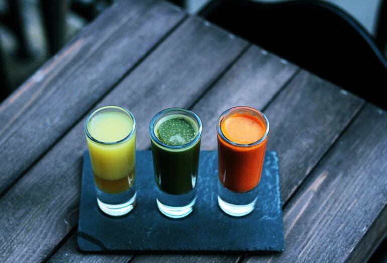 Three shot glasses full of colorful liquids sitting on a wooden bench.
