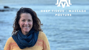 A woman in a yellow sweater and scarf stands by the ocean, with waves gently lapping at the shore in the background.