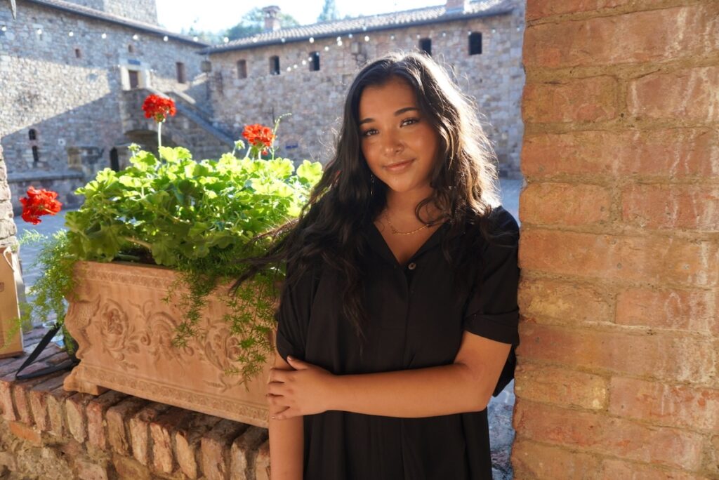 A girl with dark hair and a black dress stands next to a brick column and flower bed.