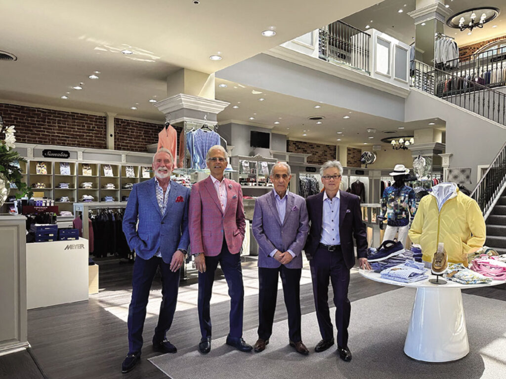 Four men stand in front of a clothing store, showcasing various garments displayed behind them.
