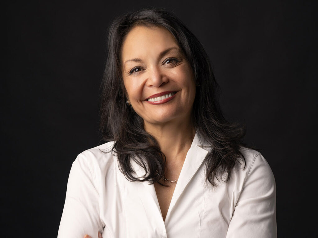 A woman with long dark hair dressed in a white shirt, looking directly at the viewer with a friendly expression.