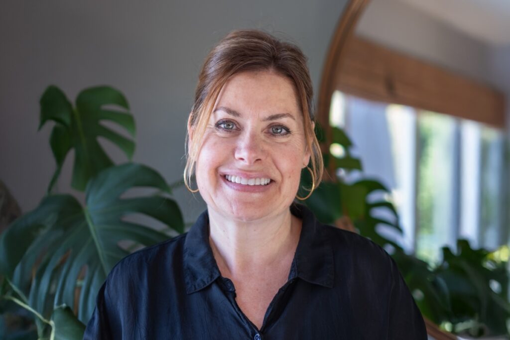 A woman wearing a black blouse and gold hoops smiles in front of a large plant and mirror.