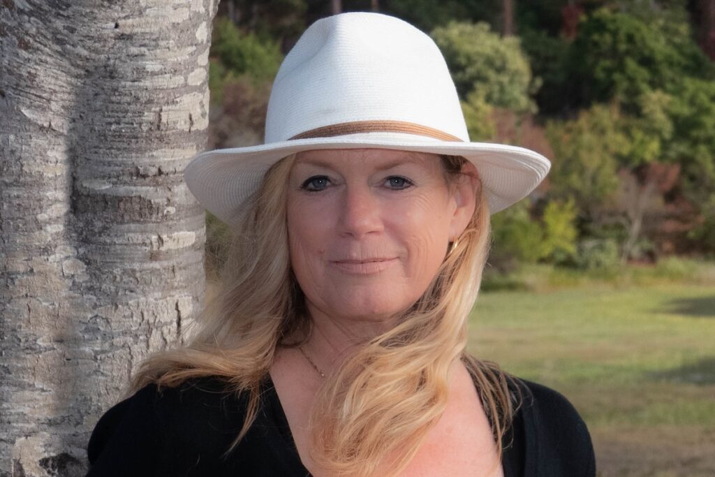 A woman with blonde hair and white brimmed hat standing next to a tree.