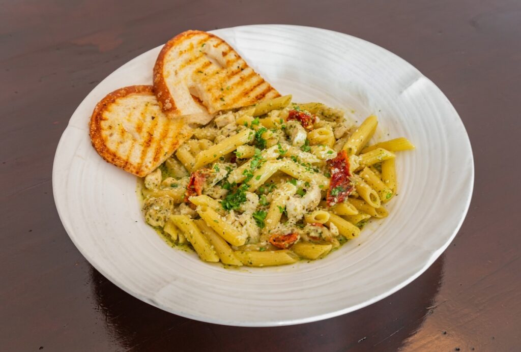 A bowl of penne pasta topped with parm, herbs and sundried tomatoes, with a side of toasted bread.
