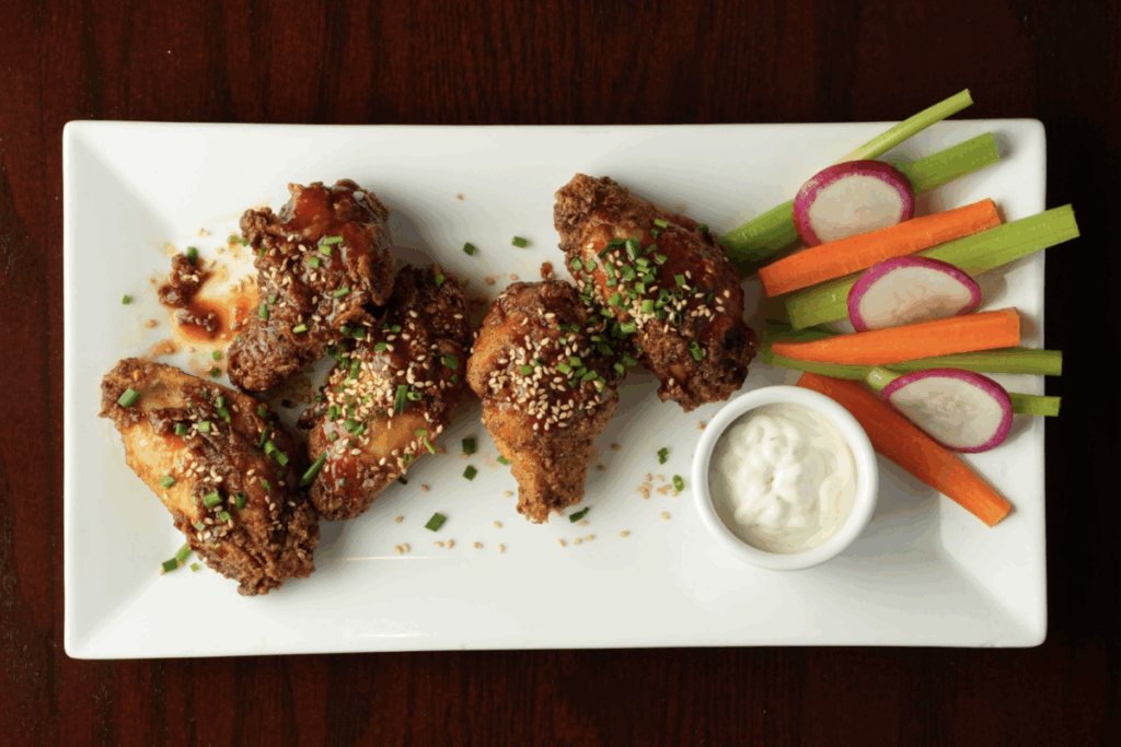 A plate with sauce-coated wings, some veggies and a white dip.