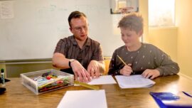 A boy and his tutor sit at a table looking at papers.