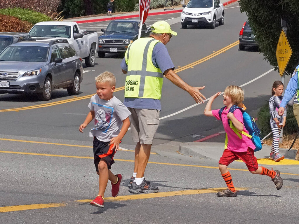 A crossing guard high-fives a smiling child with a backpack while other kids run across the street.