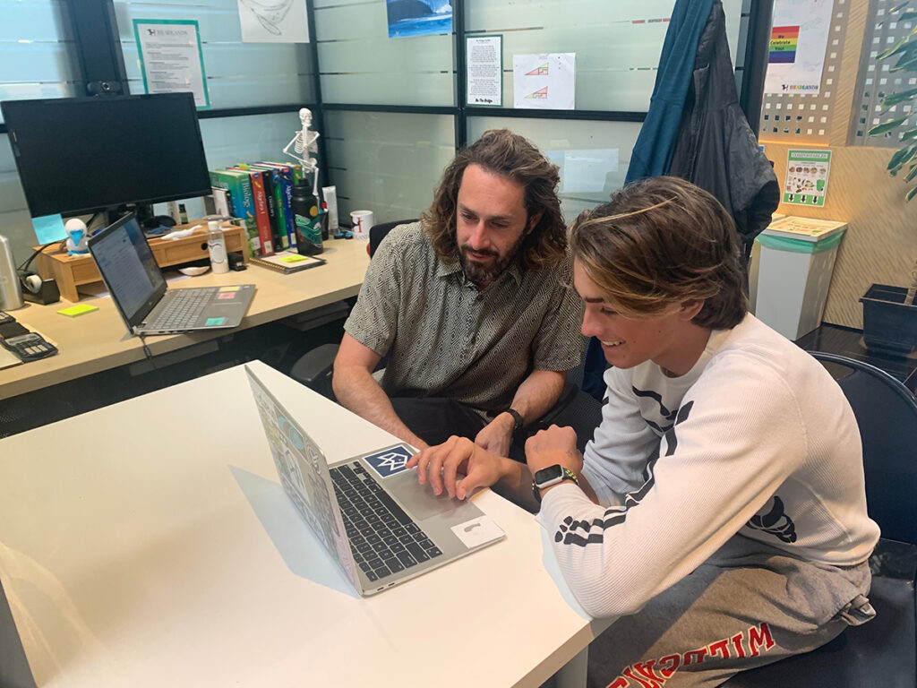 Two men seated at a desk, focused on a laptop screen, engaged in discussion or collaboration.
