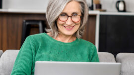 Smiling older woman with gray hair and glasses wearing a green sweater, sitting on a couch and using a laptop in a modern home setting.