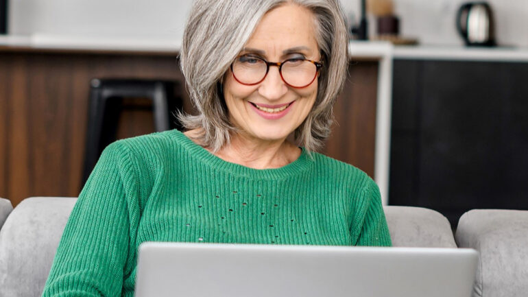Smiling older woman with gray hair and glasses wearing a green sweater, sitting on a couch and using a laptop in a modern home setting.