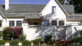 Two women sit smiling on an orange blanket on the lawn in front of a large white house with gray roof shingles, surrounded by lush greenery and purple flowering plants.