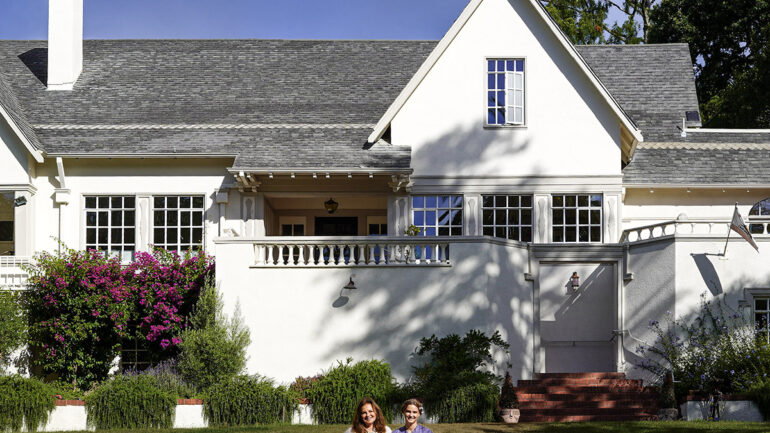 Two women sit smiling on an orange blanket on the lawn in front of a large white house with gray roof shingles, surrounded by lush greenery and purple flowering plants.