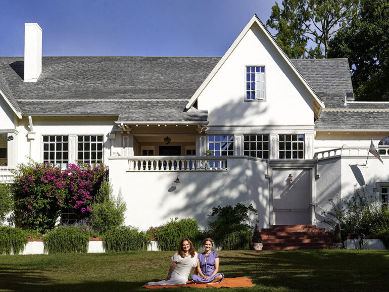 Two women sit smiling on an orange blanket on the lawn in front of a large white house with gray roof shingles, surrounded by lush greenery and purple flowering plants.