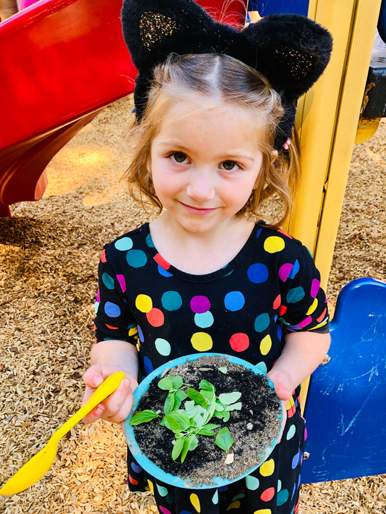 A little girl joyfully holds a bowl filled with various green plants, smiling at the camera.