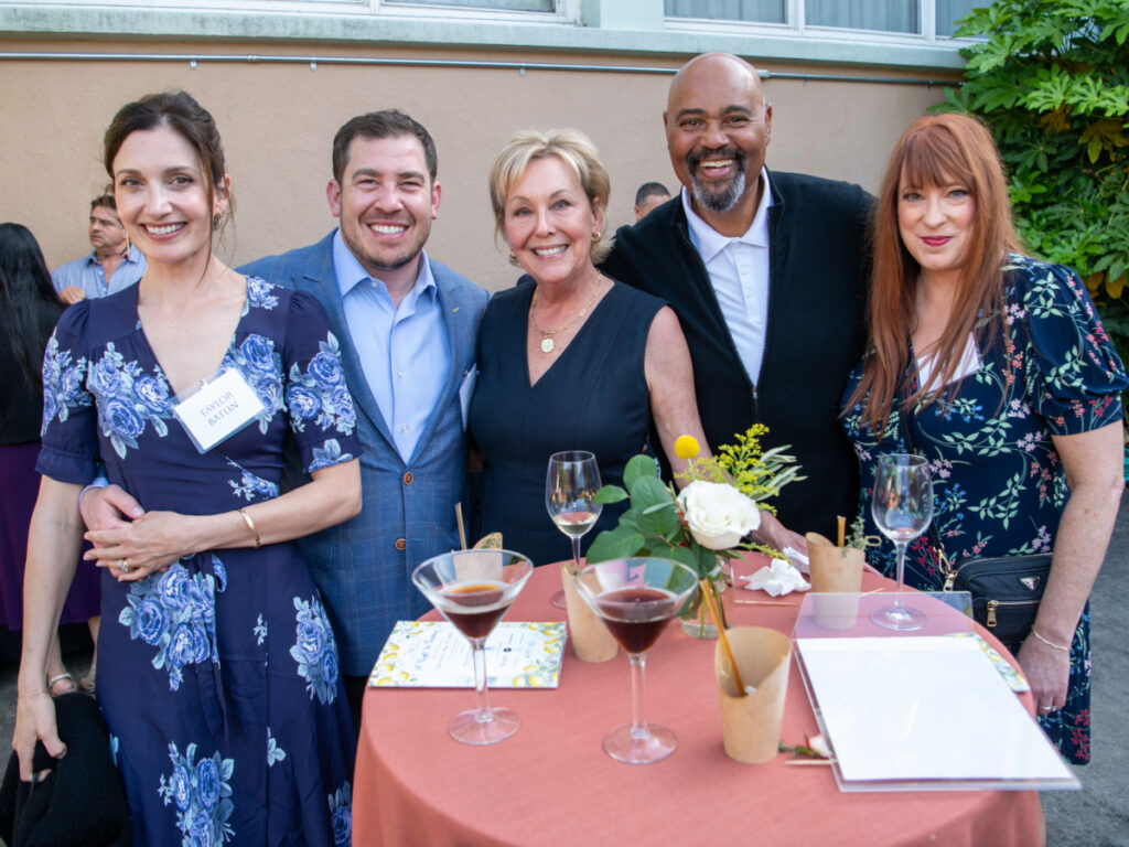 A group of five guests gather around a cocktail table outdoors, enjoying wine and conversation under the evening light at NBCC’s A Night in Tuscany benefit.